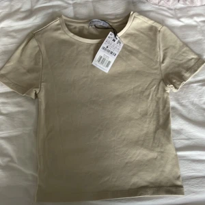 Beige t-shirt från Stradivarius - Säljer en stilren beige t-shirt från Stradivarius. Den är kortärmad och tillverkad i mjuk bomull, perfekt för en avslappnad look. Passar bra till både jeans och kjol. Ny med taggar kvar!