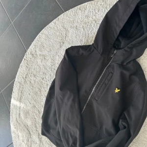 Svart jacka med huva från Lyle & Scott - Säljer en stilren svart jacka med huva från Lyle & Scott. Jackan har en dragkedja framtill och ett litet gult broderat emblem på bröstet. Perfekt för svala dagar.