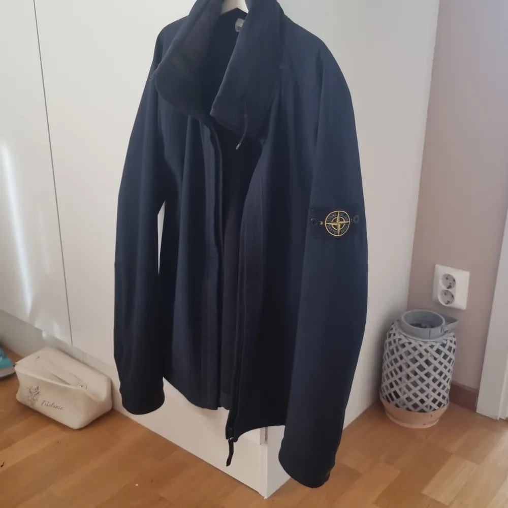 Säljer en blå jacka från Stone Island med dragkedja och långärmad design. Jackan har en broderad logotyp på ärmen och är tillverkad i ett lätt och mjukt material som är vattenavvisande.. Takit.