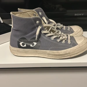 Grå sneakers från Converse x Comme des Garçons - Säljer ett par grå höga sneakers från Converse i samarbete med Comme des Garçons. Skorna har det ikoniska hjärtmotivet på sidan och klassisk snörning. Perfekta för en stilren look.