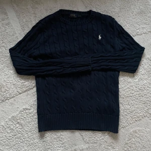 Mörkblå stickad tröja från Ralph Lauren - Säljer en snygg mörkblå stickad tröja från Ralph Lauren med det klassiska logotypbroderiet på bröstet. Tröjan har en kabelstickad design och rund halsringning.krympte 