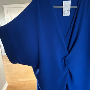 Blå klänning från Cosy House - Snygg blå klänning från Cosy House med en omlott-design och korta ärmar. Perfekt för en stilren look.