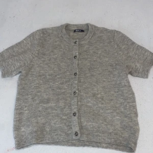 Grå stickad kofta  - Mysig grå stickad kofta med knappar framtill. Perfekt för kyliga dagar med sin kortärmade design och ribbade kanter. En klassisk och bekväm tröja för alla tillfällen.