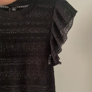 Svart topp med volangärmar - Snygg svart topp med mjukt material och volangdetaljer på ärmarna. Perfekt för en kväll ute eller en festlig tillställning. Toppen har en elegant och stilren design som passar till många olika outfits.