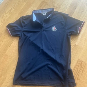 Mörkblå Moncler  - Snygg mörkblå t-shirt från Moncler med klassisk polokrage och korta ärmar. T-shirten har Moncler-logga på bröstet och coola rödvita detaljer på krage och ärmslut. Tillverkad i mjuk bomull som känns skön mot huden. Perfekt för dig som gillar stilrena märkesplagg.