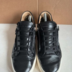 Svarta sneakers från Giuseppe Zanotti - Snygga svarta sneakers från Giuseppe Zanotti med vit platt sula och coola dragkedjor på sidorna. Skorna är gjorda i äkta skinn och har snörning framtill. Perfekt för dig som gillar stilrena och lyxiga detaljer i din outfit.