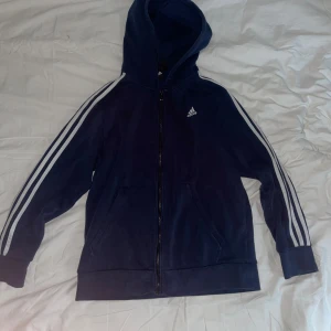 Marinblå Adidas hoodie med dragkedja - Säljer en marinblå hoodie från Adidas med klassiska vita ränder längs ärmarna och Adidas-logga på bröstet. Tröjan har huva, fickor fram och hel dragkedja. Mjuk insida i fleece och ribbade muddar vid ärmslut och nederkant. Perfekt för chill eller sport.