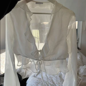 Vit omlottblus med volang från Bershka - Superfin vit omlottblus från Bershka i transparent, lätt material. Blusen har långa ärmar, volangkant längs urringningen och knyt framtill i midjan. Perfekt för lager-på-lager och ger en romantisk vibe till din outfit.
