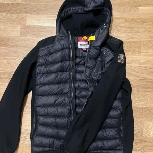 Parajumper jacka - Svart pufferjacka från Parajumpers med huva och coolt gult band vid kragen. Jackan har quiltad framsida och rygg, samt släta svarta ärmar med PJS-logga på axeln. Dragkedja framtill och sidofickor. Perfekt för kyliga dagar.
