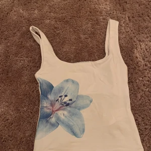 Vit linnetopp med blå blomma Subdued - Supersöt vit linnetopp från Subdued i storlek XS. Toppen har breda axelband och en stor blå blomma tryckt framtill som ger en fräsch och somrig vibe. Perfekt att styla med jeans eller kjol för en chill look.