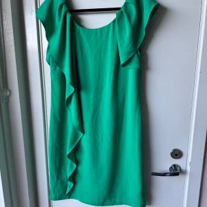 Grön kortklänning med volang från Vila - Snygg grön klänning från Vila i storlek S. Modellen är kort med volangdetalj längs ena sidan och har en rundad halsringning. Klänningen har korta ärmar och är tillverkad i ett mjukt, lätt polyestertyg som ger ett skönt fall. Perfekt för dig som gillar färg och vill sticka ut.