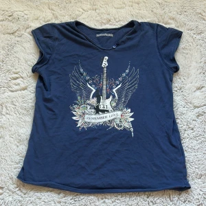 Blå topp Zadig & Voltaire med tryck - Cool blå topp från Zadig & Voltaire med korta ärmar och rund hals med två knappar. Framsidan har ett tryck med elgitarr, vingar och blommor samt texten 'Remember Love'. Perfekt för dig som gillar rockig stil och snygga detaljer. Bra skick och storlek 152 men passar 158 samt xs
