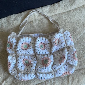 Handgjord blommig stickad axelväska - Superfin axelväska som är stickad i ett blommigt mönster med vita, beige, gröna och ljusrosa färger. Väskan har två handtag och är gjord av mjukt garn, perfekt för att bära dina grejer på ett unikt sätt. Blommorna är virkade i rutmönster och ger en riktigt mysig vibe.