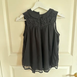 Svart blus med spets från KappAhl - Supersöt svart blus från KappAhl med spets upptill och klassisk krage. Ärmlös modell med knappar framtill och lätt, luftig känsla. Perfekt när du vill ha något stilrent men ändå med fina detaljer.