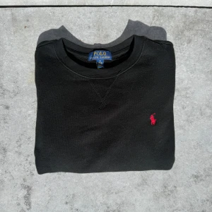 Svart tröja från Polo Ralph Lauren - Svart tröja från Polo Ralph Lauren med klassisk rund hals och röd broderad logga på bröstet. Tröjan är i mjuk bomull och har ribbade muddar vid hals och ärmslut. Perfekt för dig som gillar stilrena och tidlösa plagg.