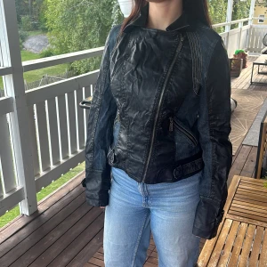 Vintage❤️ - Cool bikerinspirerad jacka i svart skinn och blå denim med sned dragkedja, markerade sömmar och detaljer vid ärmslut och midja. Jackan har lång ärm och en figurnära passform som ger en edgy look. Perfekt för dig som vill sticka ut med en unik stil. En defekt se sista bilden. Vintage