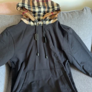 Svart Burberry hoodie med rutig huva - Säljer en svart hoodie från Burberry i storlek L. Den har en klassisk rutig huva i beige, svart, vit och röd, samt snörning med Burberry-logga. Tröjan har en stor magficka och är tillverkad i mjuk bomull. Perfekt för dig som gillar stilrena streetwear-plagg med lyxig touch.