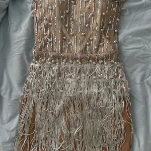 Champagnefärgad paljettklänning Bella Barnett - Supersnygg kort klänning i champagne med glittriga paljetter, pärlor och långa fransar. Märket är Bella Barnett. Klänningen har en figurnära passform och är perfekt för dig som vill sticka ut. Materialet är polyester med transparent mesh och detaljer i silver.