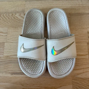 Vita Nike sandaler med holografisk logo - Säljer ett par vita Nike sandaler med bred rem och holografisk Swoosh-logga framtill. Skorna har en strukturerad innersula för extra komfort och en platt yttersula med Nike-mönster. Perfekta för chill dagar och enkel att slänga på sig. Materialet är syntet och färgen är vit med metallic-detalj.