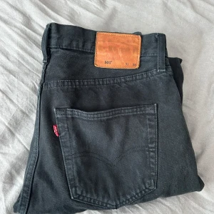 Svarta Levi's 501 jeans med rå kant - Svarta Levi's 501 jeans med klassisk och röd Levi's-tag på bakfickan. Jeansen har rak passform och är tillverkade i slitstarkt bomullsjeansmaterial. Nedre delen av benen har en rå, fransad kant som ger en cool och avslappnad vibe.