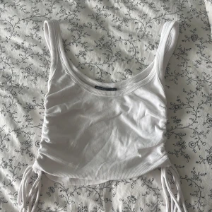 Vit croppad linnetopp från Zara - Supersnygg vit croppad linnetopp från Zara med ribbad struktur och snörning på sidorna. Toppen har breda axelband och är ärmlös, perfekt för att styla med jeans eller kjol. Materialet är mjuk bomull med lite stretch för skön passform.