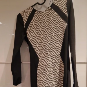 Svart och vit mönstrad långärmad klänning - Snygg långärmad klänning med svart bas och vit geometriskt mönstrad panel framtill. Klänningen har en smickrande form med svarta sidor och rund halsringning. Perfekt för dig som gillar grafiska detaljer och stilren design.