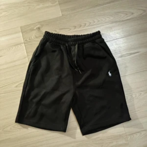 Svarta shorts från Polo Ralph Lauren - Svarta shorts från Polo Ralph Lauren med elastisk midja och snörning. Klassisk design med två sidofickor och broderad logga på vänster ben. Perfekta för chill dagar eller träning, tillverkade i mjuk bomull för extra komfort.