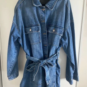 Blå jeansklänning med bälte - Säljer en cool blå jeansklänning med långa ärmar och knappar framtill. Klänningen har två stora bröstfickor med knappstängning och ett matchande bälte i midjan för en snygg siluett. Perfekt för dig som gillar avslappnad denimstil.