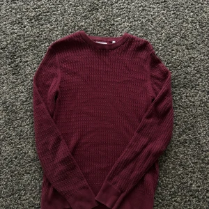 Vinröd stickad tröja från Bläck - Säljer en vinröd stickad tröja från Bläck med snyggt strukturerat mönster. Tröjan har rund hals och långa ärmar, perfekt för dig som gillar stilrena och enkla plagg med lite extra textur. Passar dig som vill ha en klassisk look med en twist.