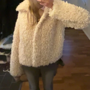Beige fluffig teddyjacka - Supermysig teddyjacka i beige med fluffig och krullig yta. Jackan har en bred krage och långärmade ärmar, perfekt för dig som gillar en chill och trendig look. Passformen är loose och jackan är tillverkad i syntetmaterial som ger en skön känsla. Kom privat för fler bilder! 