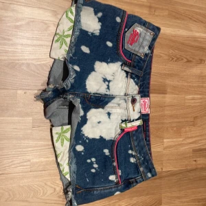 Superdry jeansshorts med blekta detaljer - Säljer ett par Superdry jeansshorts med coolt blekt mönster och råa kanter. Shortsen har rosa sömmar, gröna blad på insidan av benen och flera fickor. De är lågmidjade och har en loose passform, perfekt för sommarens chill dagar. Super unika och fina