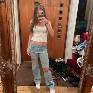 Ljusblå bootcut jeans med slitningar - Säljer ett par ljusblå jeans med bootcut passform och låg midja. Jeansen har snygga slitningar och hål på låren och knäet för en cool, avslappnad vibe. Klassisk femficksmodell med dragkedja och knapp. Perfekt för dig som gillar streetstyle.