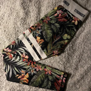 Stronger leggings med tropiskt mönster - Snygga leggings från Stronger i storlek small med tropiskt blommigt mönster i grönt, gult, rosa och rött. De har vit midja med logga och vita ränder på ena benet. Dem stretchiga och bekväma för träning. 🩷🩷