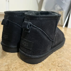 Svarta UGG boots i mocka, storlek 44 - Svarta UGG boots med rund tå och platt sula. Utsidan är i mjuk mocka och insidan är fodrad med fluffigt syntetmaterial för extra värme. Klassisk UGG-logga baktill och grovt mönstrad yttersula. Perfekt för kalla dagar och chill stil.
