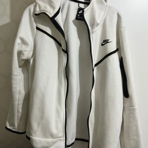 Vit Nike jacka med svarta detaljer - Snygg vit jacka från Nike i storlek S med svarta kontrastdetaljer och dragkedja framtill. Jackan har lång ärm och en cool ficka med dragkedja på ena ärmen. Perfekt för dig som gillar sportig och clean stil.