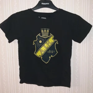 Svart AIK t-shirt med tryck - Svart t-shirt från AIK Official Merchandise med stort AIK-emblem i gult och mörkblått på bröstet. Klassisk rund hals och korta ärmar. Tillverkad i 100% bomull för skön och mjuk känsla. Perfekt för dig som vill visa ditt AIK-supporterhjärta.