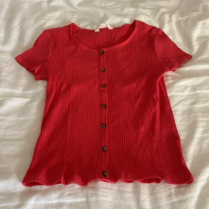 Röd ribbad topp med knappar H&M - Snygg röd ribbad topp från H&M med korta ärmar och knappar hela vägen ned. Perfekt för dig som gillar enkel men trendig stil. 