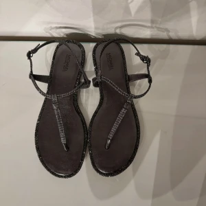 Svart/gråa sandaler Michael Kors | Nypris 1200kr - Snygga svarta sandaler från Michael Kors med glittriga remmar och platt sula. Tåremmen och de tunna remmarna runt foten ger en stilren look, perfekt för dig som gillar att sticka ut med lite extra bling. Spänne vid ankeln för justerbar passform.