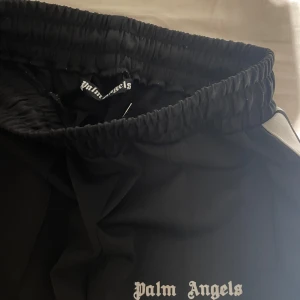 Palm Angels svarta shorts med vita ränder - Svarta shorts från Palm Angels med breda vita ränder längs sidorna och logotyp broderad framtill. Modellen har elastisk midja och två sidofickor. Materialet är mjukt och har en lätt glans, perfekt för en chill streetwear-look.