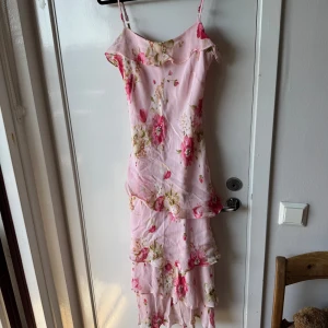 Ljusrosa blommig långklänning American Angel - Superfin ljusrosa långklänning från American Angel med blommigt mönster i rosa och beige toner. Klänningen har smala axelband, volangdetaljer och flera lager i kjolen. Materialet är rayon med polyesterfoder, perfekt för sommarens alla tillfällen.