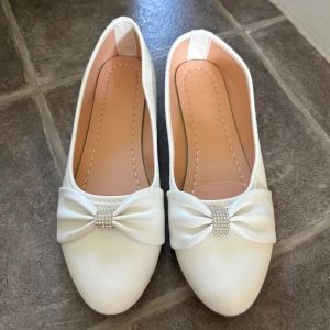 Vita ballerinaskor med rosett och strass - Snygga vita ballerinaskor från SITAYT med rund tå och dekorativ rosett framtill, prydd med glittrande strassdetaljer. Skorna har en platt sula och är tillverkade i syntetmaterial med mjuk innersula för extra komfort. Är i storlek 40 men är små i storleken så jag skulle säga att de passar en 38