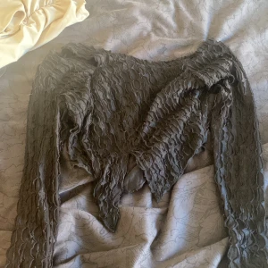 Svart omlottopp med lång ärm - Säljer en svart omlottopp med lång ärm och coolt texturerat tyg. Toppen har en v-ringad front och ett vågigt, nästan quiltat mönster som ger en unik look. Perfekt för dig som vill sticka ut med en edgy stil.