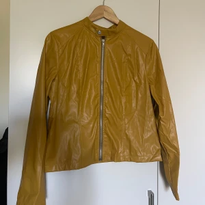 Gul skinnjacka med dragkedja - Cool gul fejkskinnjacka med rak passform och silverfärgad dragkedja framtill. Jackan har ståkrage med tryckknapp och långa ärmar. Materialet är slätt och glansigt, vilket ger en modern vibe. Perfekt för dig som vill sticka ut med färg och stil.helt ny oanvänd 