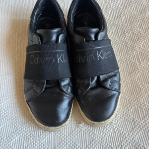 Svarta sneakers från Calvin Klein - Svarta sneakers från Calvin Klein med bred elastisk rem över foten med logotyp. Ovandel i skinn och mockadetalj vid hälen, samt vit platt sula med CK-mönster undertill. Snygg och stilren design som passar till jeans och hoodie.