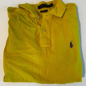 Gul pikétröja från Polo Ralph Lauren - Snygg gul pikétröja från Polo Ralph Lauren i klassisk passform. Tröjan har krage, två knappar och den ikoniska lila broderade loggan på bröstet. Materialet är bomull med en lite grov struktur som känns chill och stilrent. 