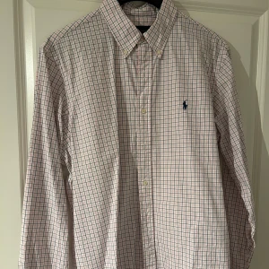 Ralph Lauren skjorta  - Klassisk rutig skjorta från Ralph Lauren – en perfekt blandning av stil och komfort! Denna skjorta är tillverkad i mjukt bomullsmaterial och har den ikoniska Ralph Lauren-loggan på bröstet. Passar lika bra till en avslappnad look med jeans eller uppklädd med chinos för en smart vardagsoutfit. Inga fläckar eller defekter – välvårdad och redo för användning!