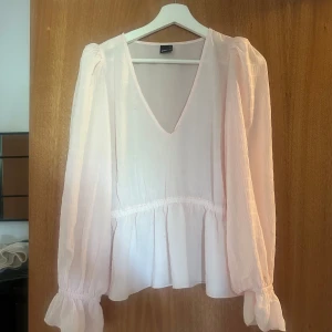 Ljusrosa blus med volang och v-ringning - Superfin ljusrosa blus från Gina Tricot med v-ringad hals och volang nedtill. Blusen har långa puffärmar med rynkade muddar och är gjord i ett lätt, luftigt material som känns skönt mot huden. Perfekt för dig som gillar romantisk och feminin stil.