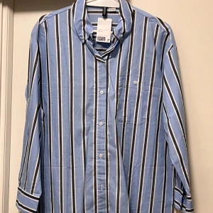 Blåvit randig skjorta från H&M Divided - Snygg blå skjorta med vita och svarta ränder från H&M Divided. Klassisk krage, bröstficka och knappar framtill. Långärmad modell i mjuk bomull som är perfekt för en chill och stilren look. Passar dig som gillar enkel men trendig stil.