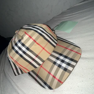 Burberry rutig keps i beige och svart - Snygg keps från Burberry med klassiskt rutigt mönster i beige, svart, vitt och röda detaljer. Justerbar baksida med svart plastspänne och Burberry London England-logga. Tillverkad i polyester, perfekt för dig som vill ha en ikonisk streetstil.