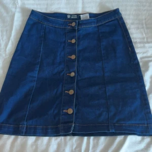 Blå jeanskjol med knappar - Snygg blå jeanskjol från Jane Skirt med raka linjer och knappar hela vägen framtill. Klassisk modell i denim som är lätt att matcha med olika toppar. Perfekt för dig som gillar en enkel men trendig stil.